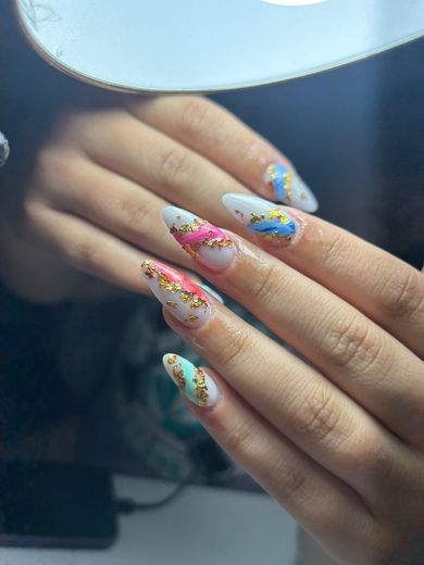 Acrílicas stiletto blancas con vetas rosa, azul, roja y verde agua y foil dorado, junto al aro de luz