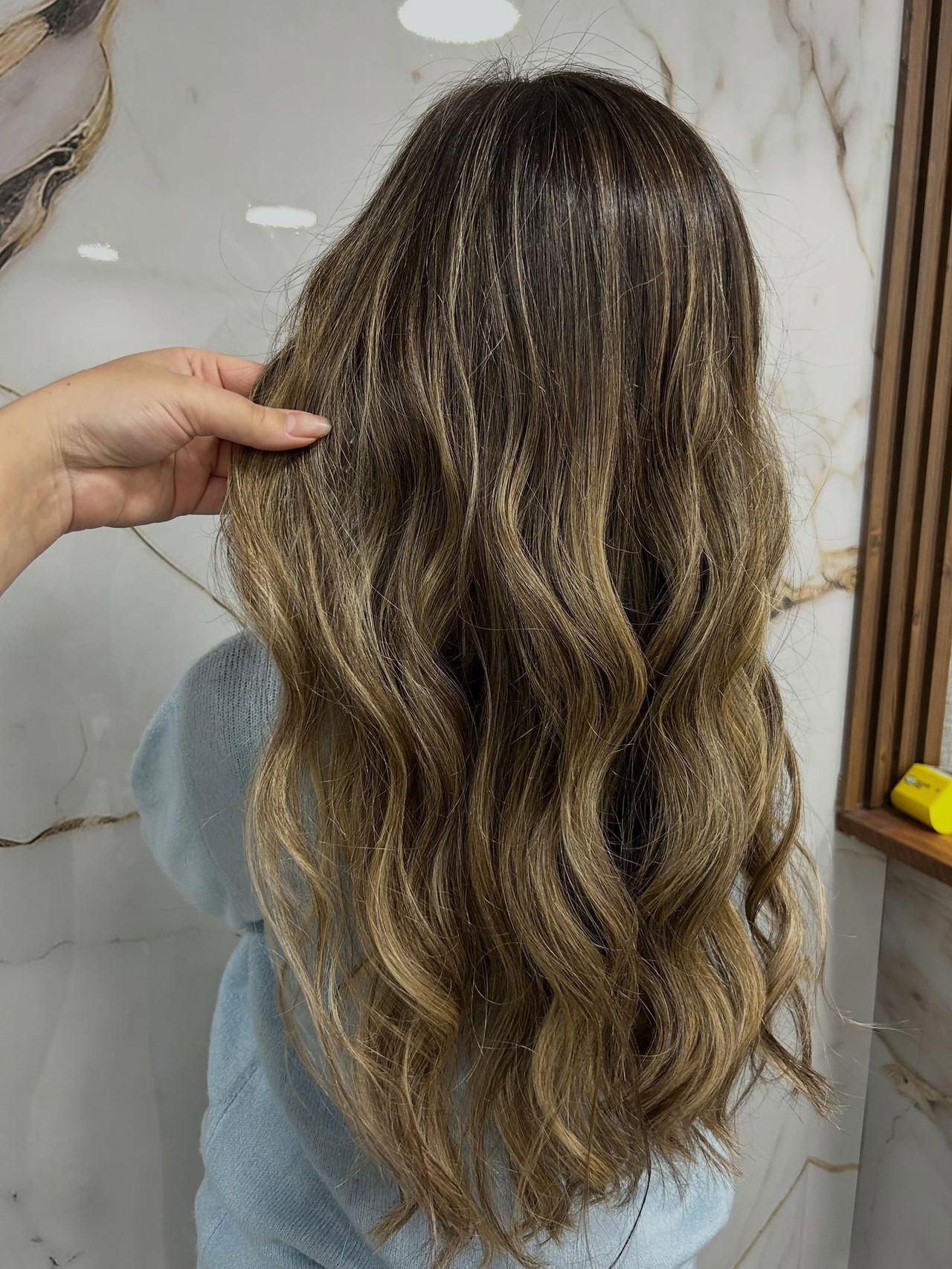Retoque de balayage: ondas largas con reflejos rubio ceniza, de espaldas contra la pared de mármol